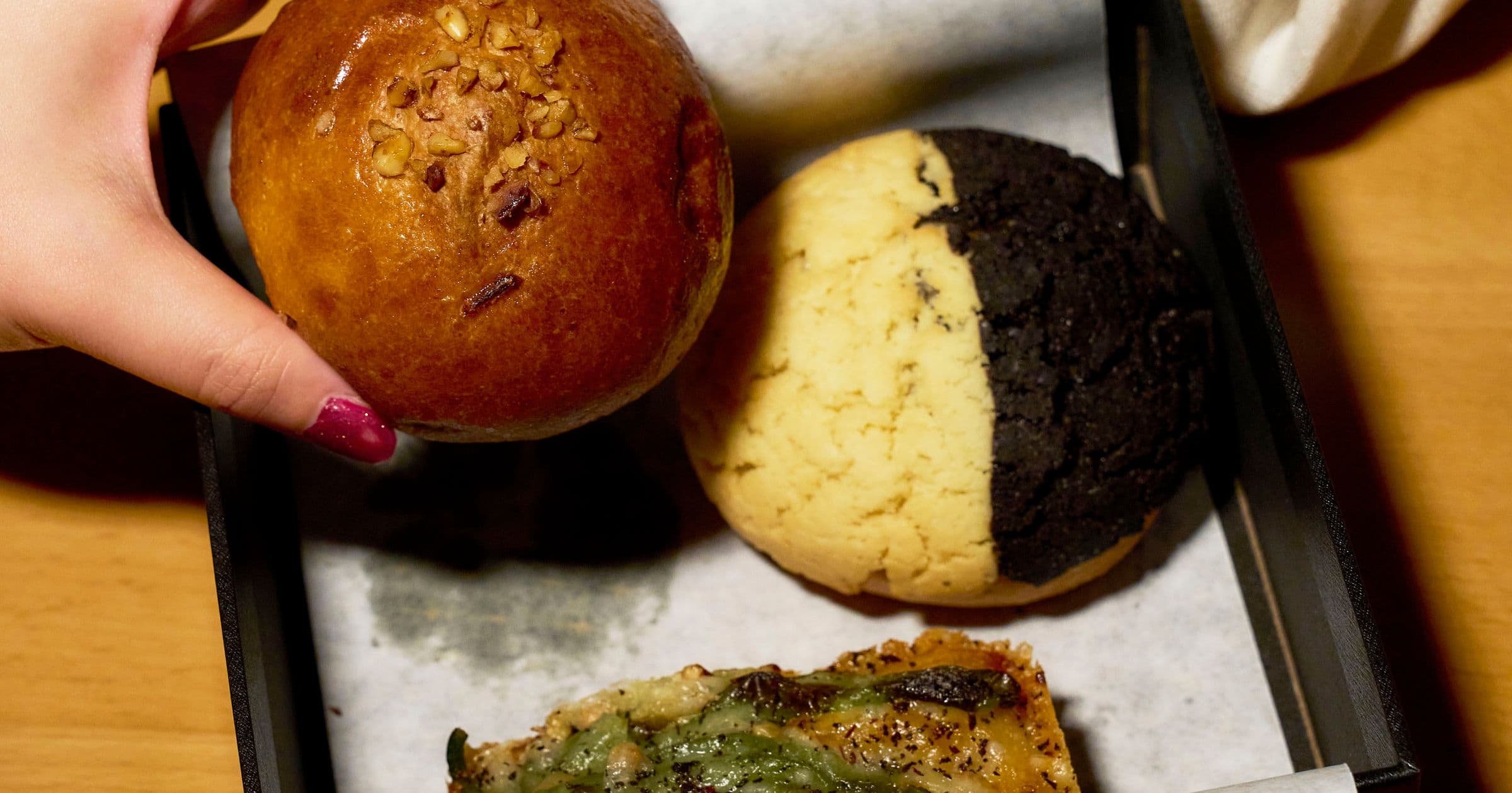 A hand holding a walnut and red-bean anpan above a black-and-white melon pan and a shishito focaccia in a black box