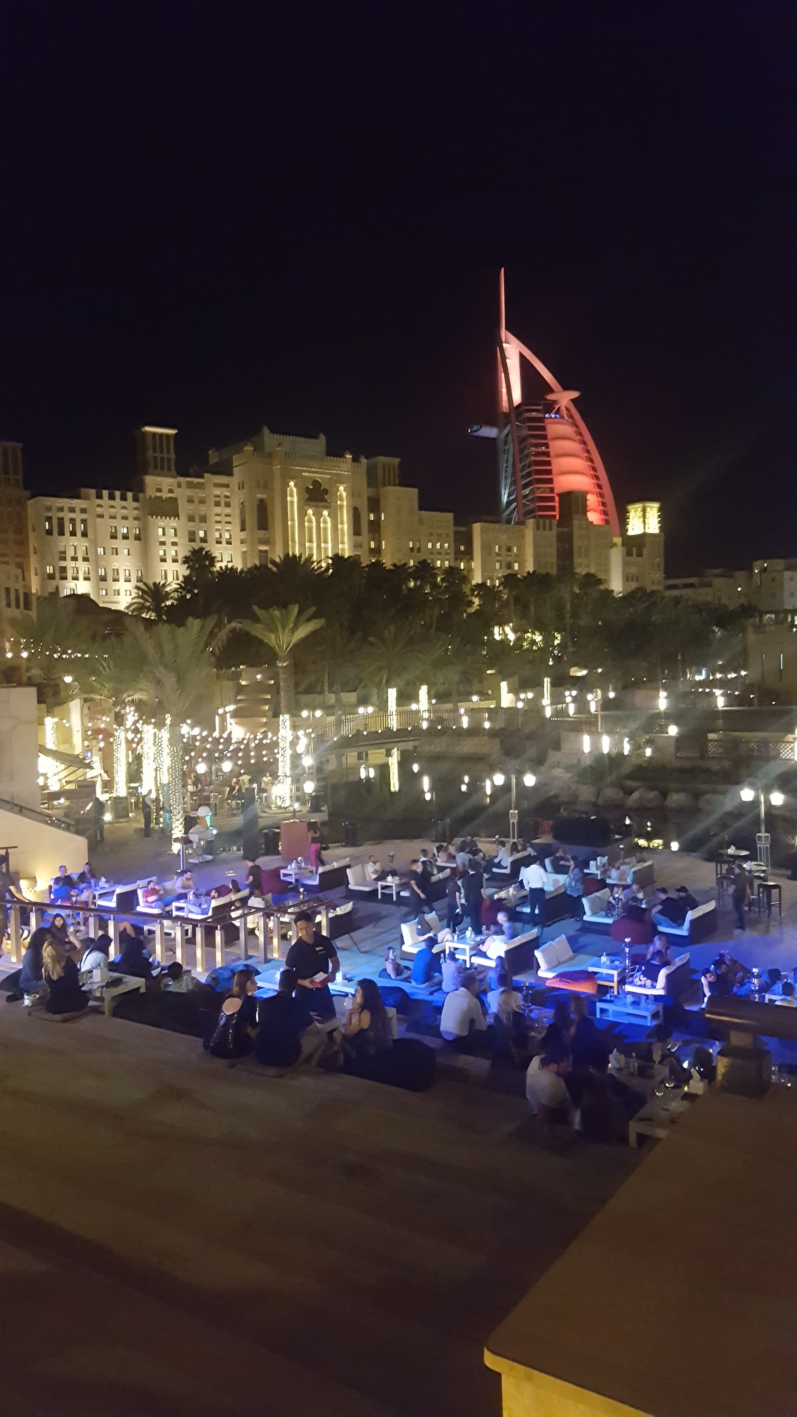 The Burj Al Arab lit red at night, with a crowded terrace and palm trees in the foreground