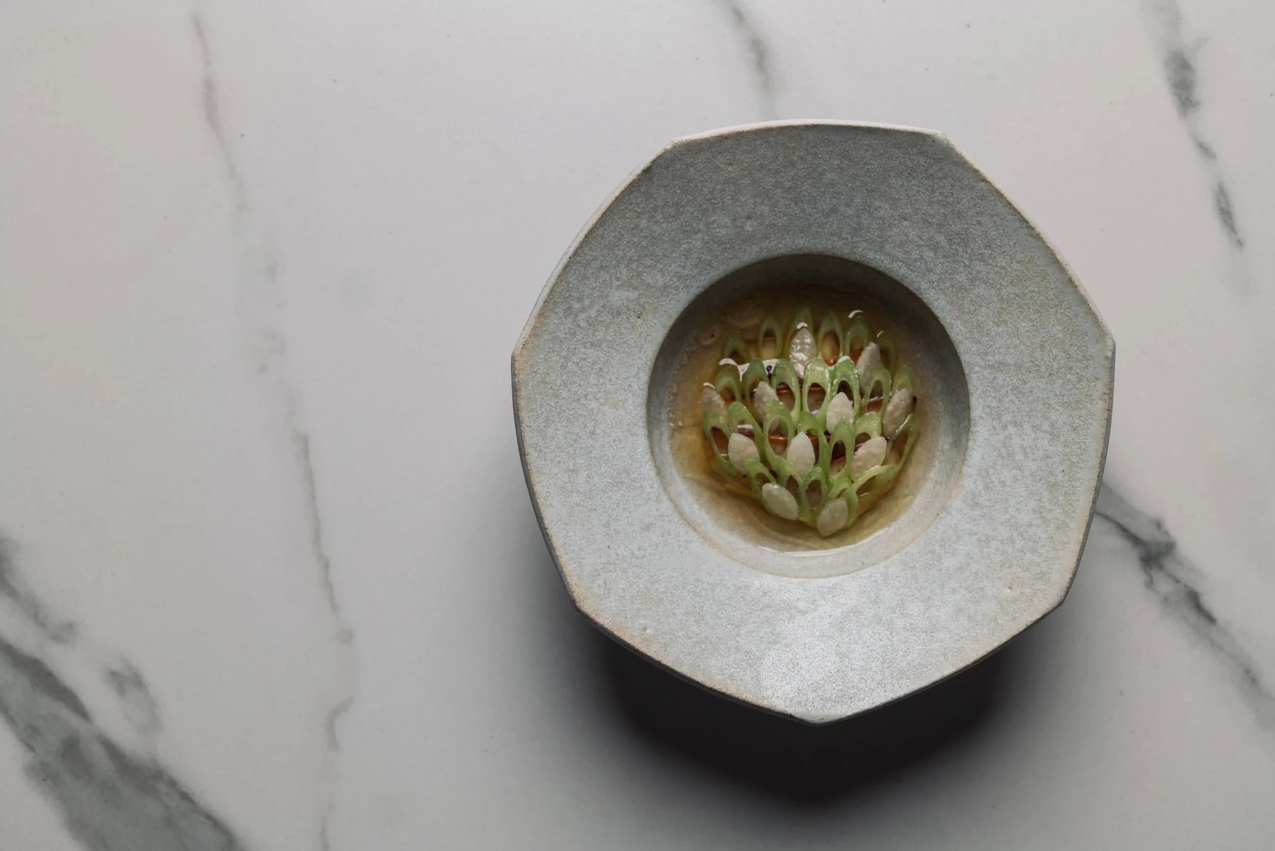 Overhead view of a sculptural octagonal ceramic bowl with a lattice of pale green slices on white marble at Restaurant Naides in San Francisco