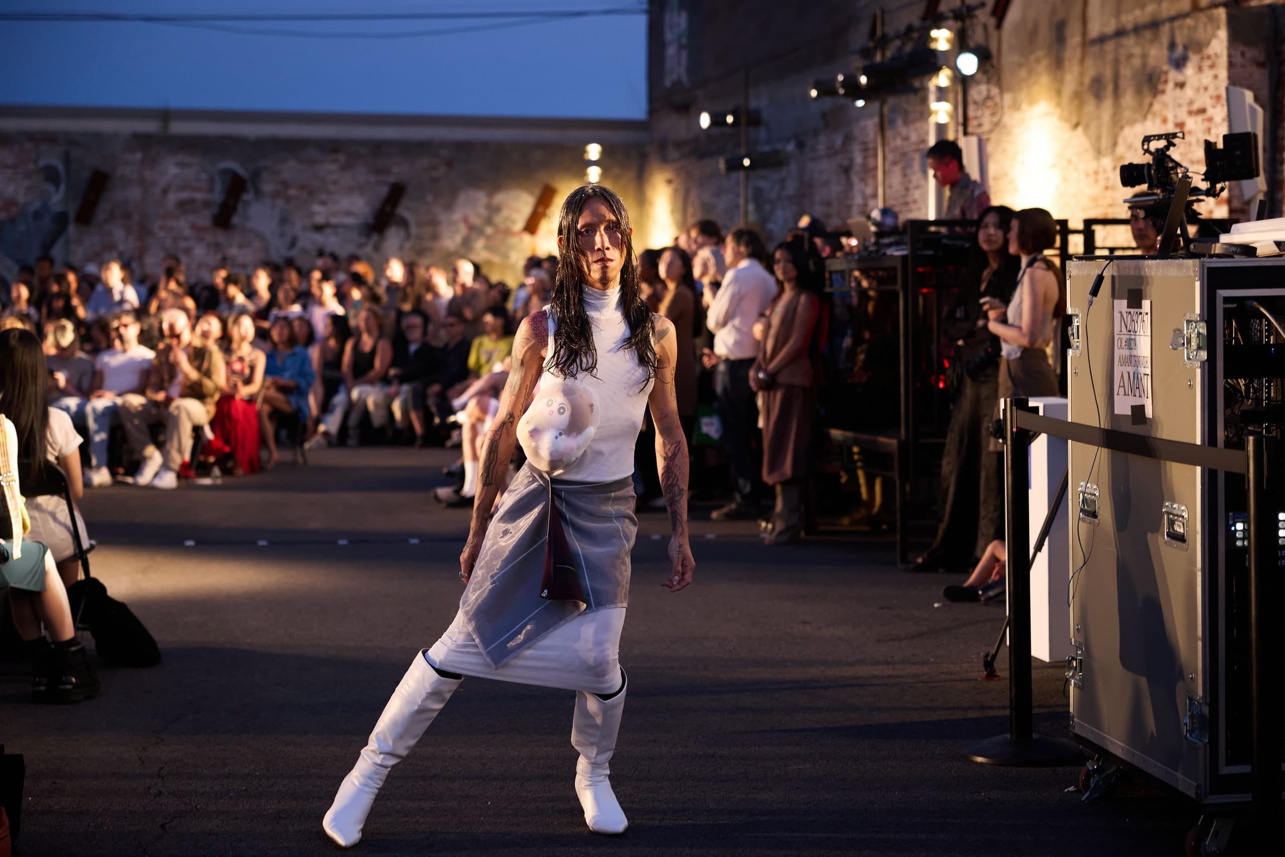 Model in a white turtleneck with a stuffed waist bump walking CFGNY’s outdoor lot runway at Amant, Brooklyn, with audience and an Amant flight case behind