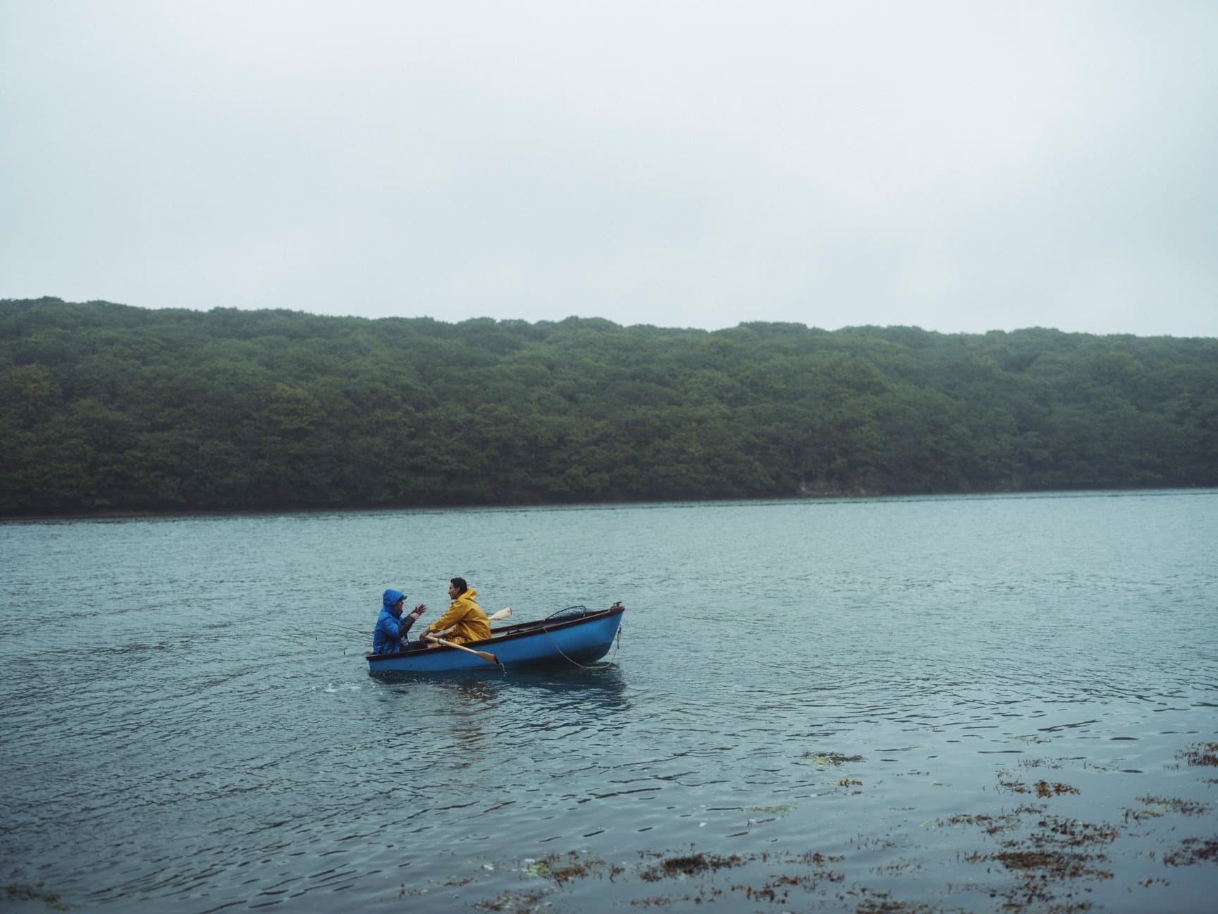 Two people in a small blue boat on a grey inlet, with a forested shore behind them