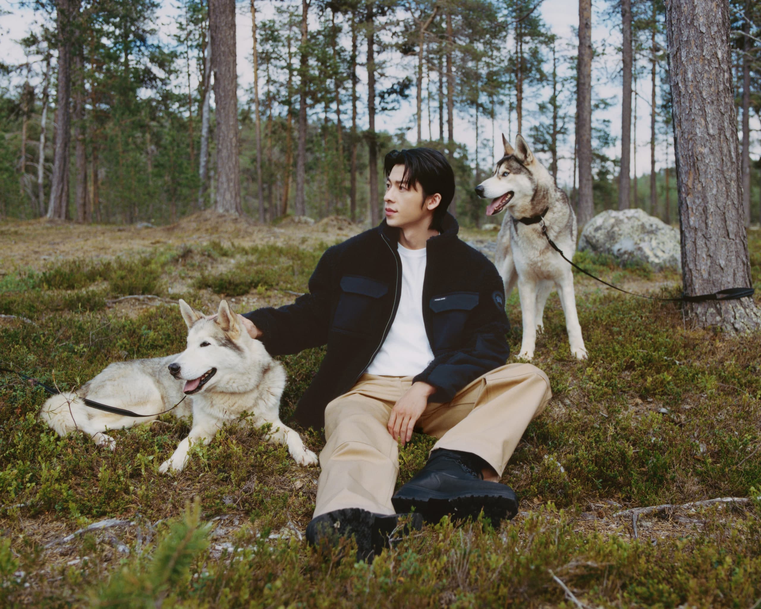 Greg Hsu seated in a Finnish pine forest with two husky dogs, wearing a black Canada Goose fleece