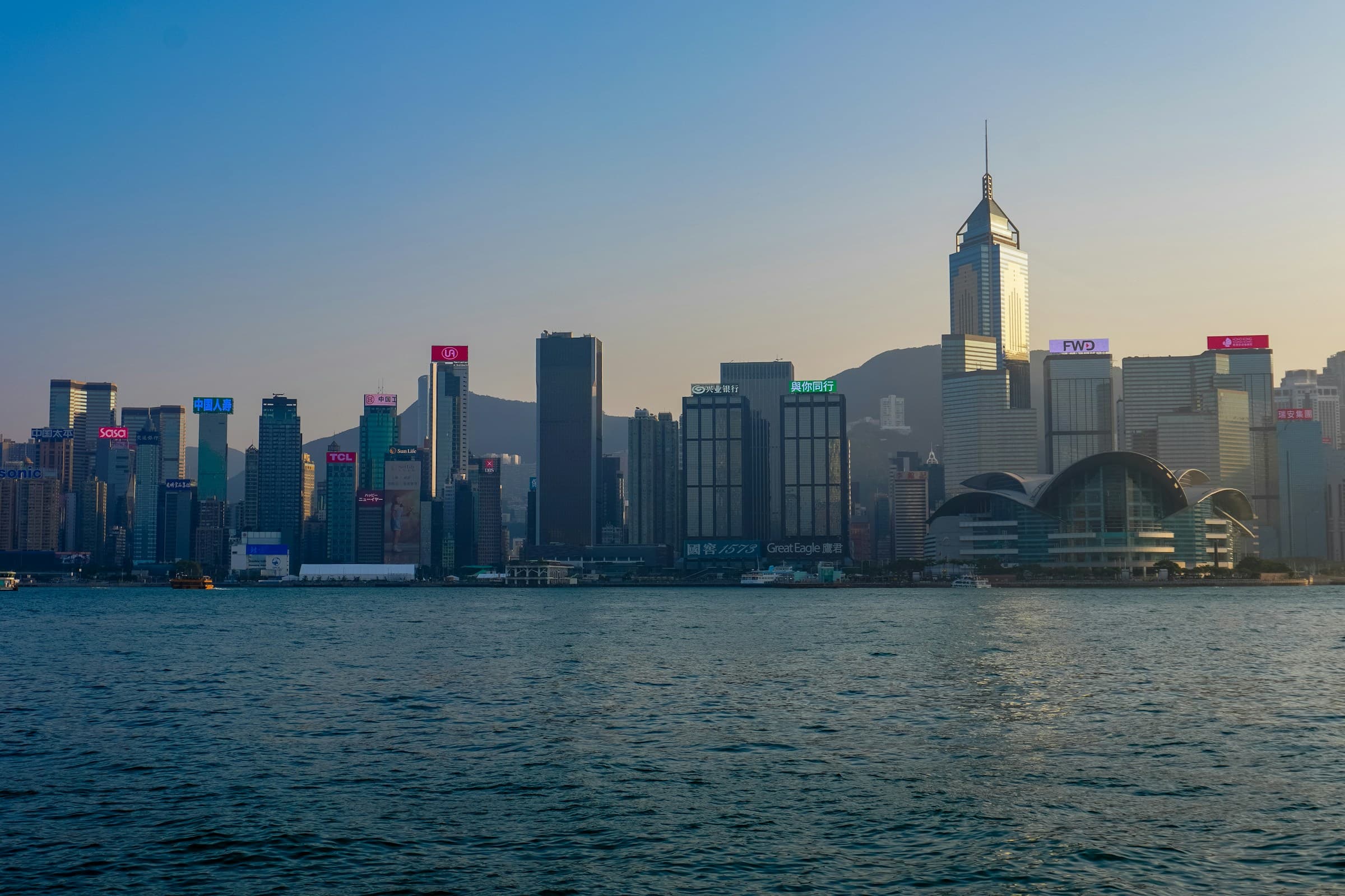 Hong Kong Island skyline in daylight across Victoria Harbour, with the Convention Centre at the right