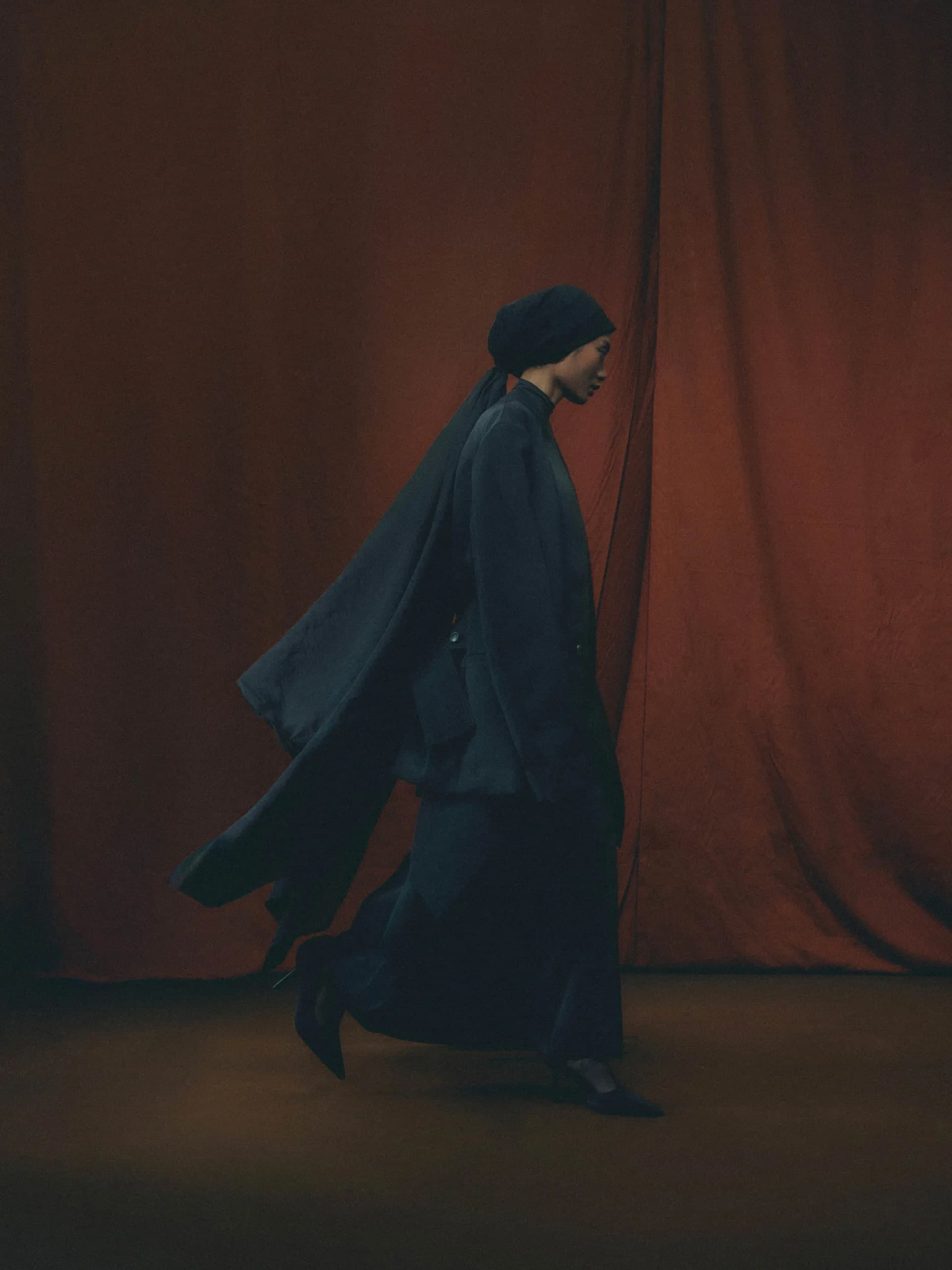 Kit Woo look: oversized funnel-neck coat, wrapped head covering, and a trailing sash against rust curtains