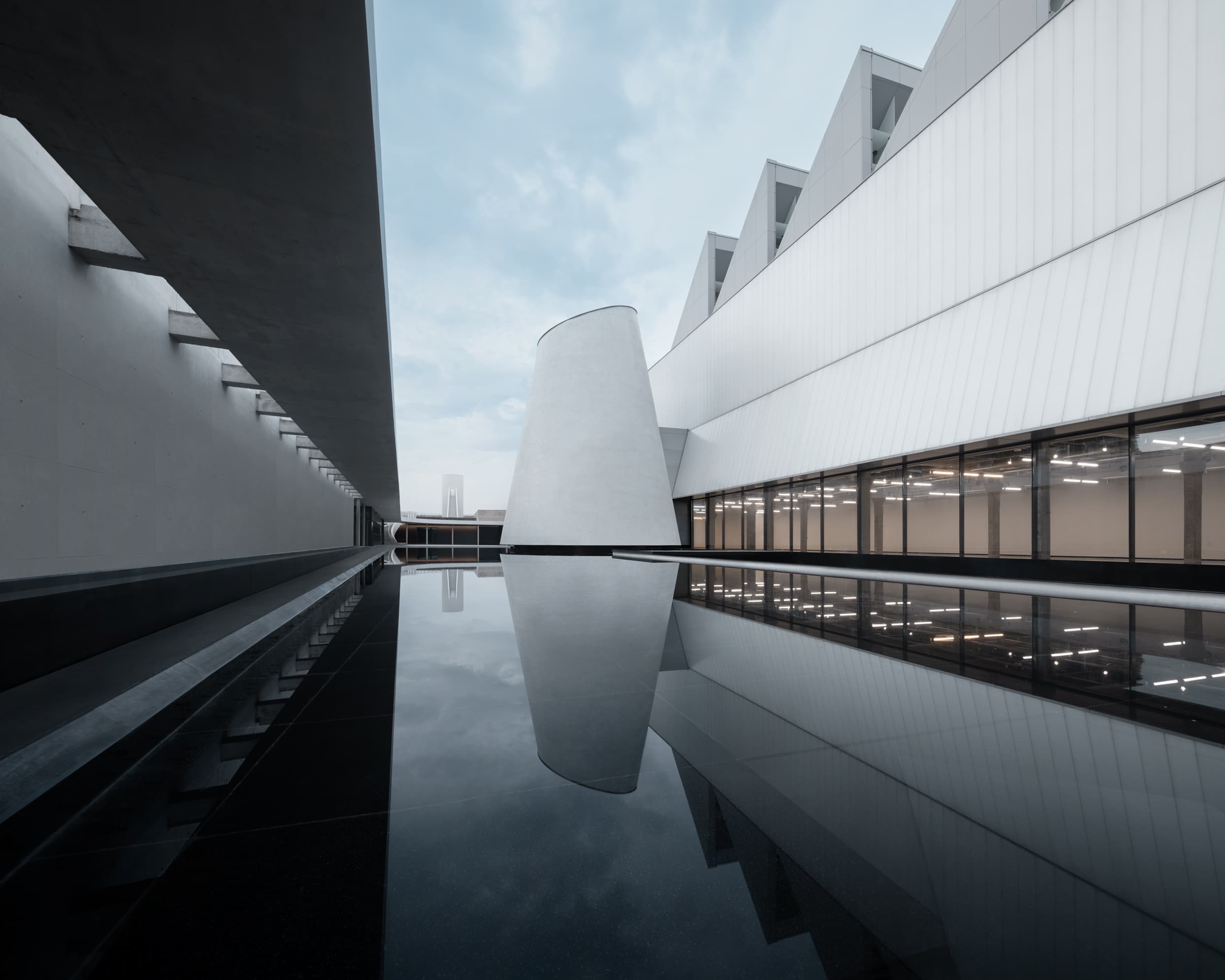 Wide view of Dib Bangkok's central courtyard, with the cone-shaped Chapel reflected in a still pool and a small figure at the far end