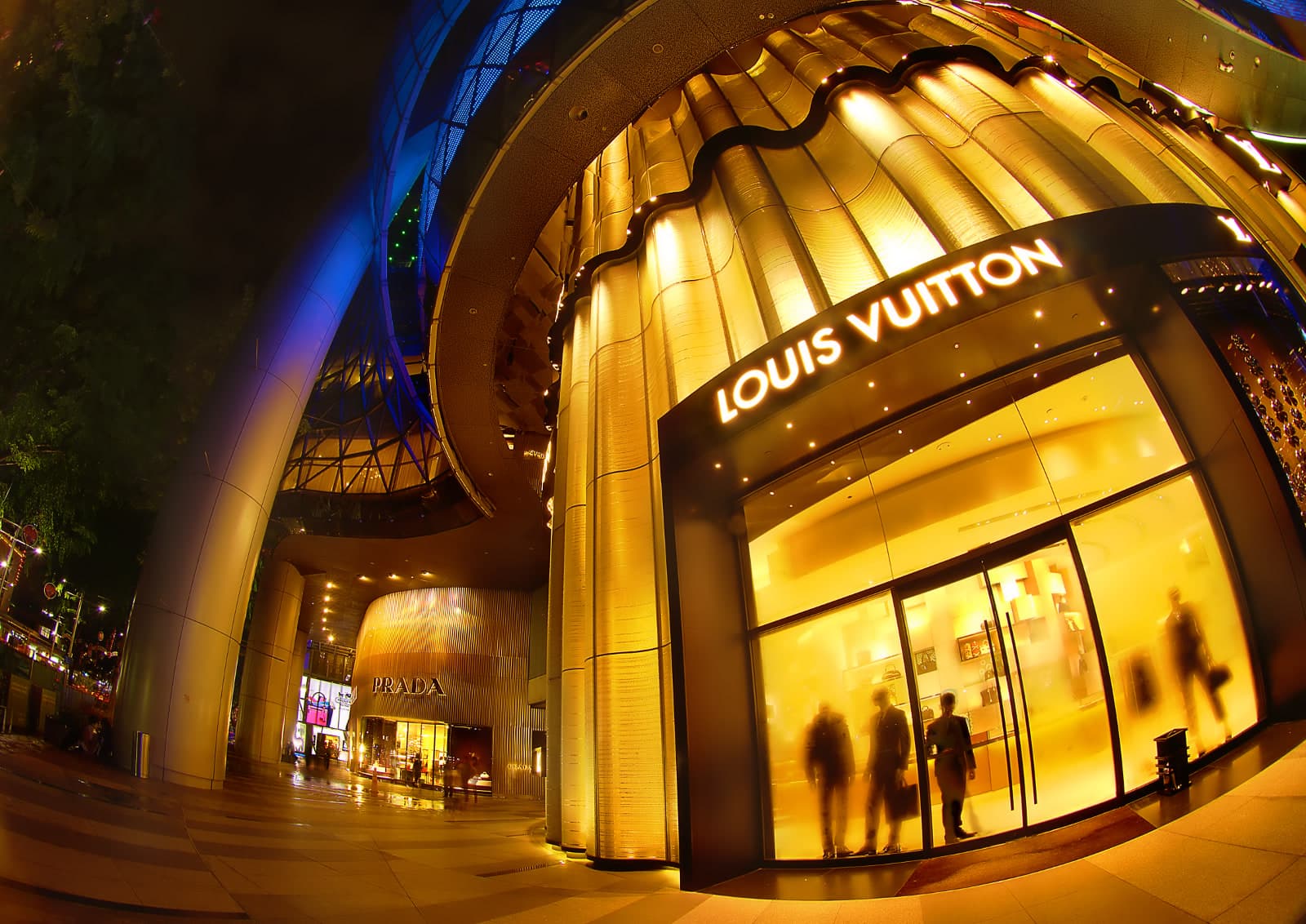 The Louis Vuitton store at ION Orchard in Singapore at night, Prada next door, shoppers visible through the glass