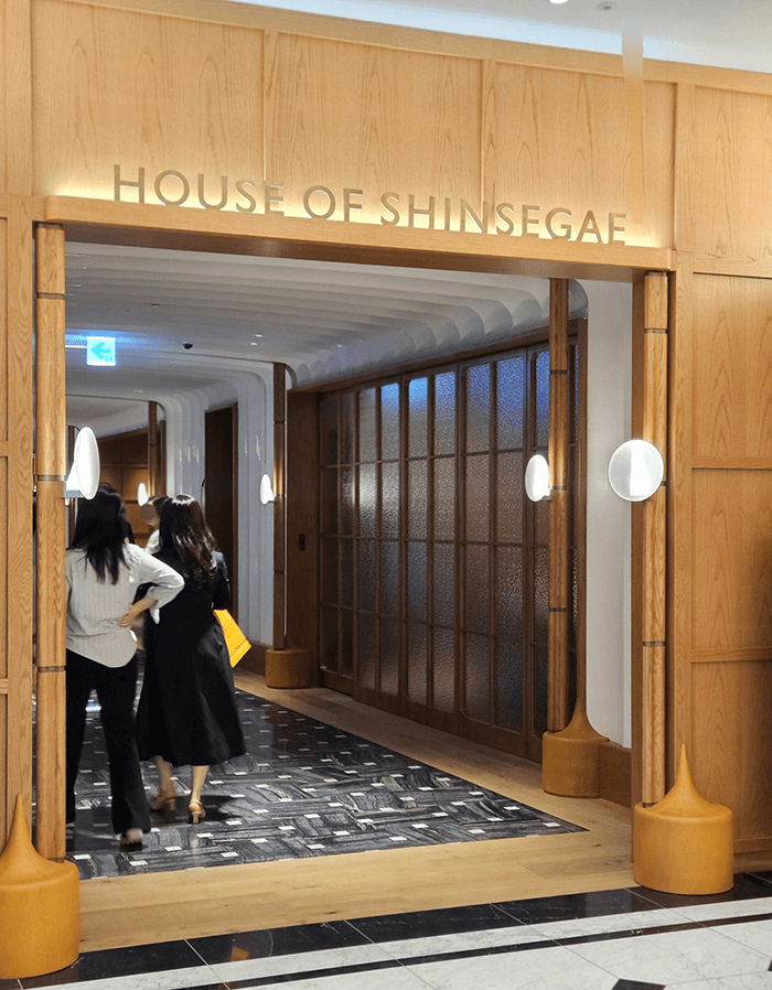 Wood-framed entrance marked House of Shinsegae, with two shoppers walking into a corridor of ribbed glass and oval wall lights