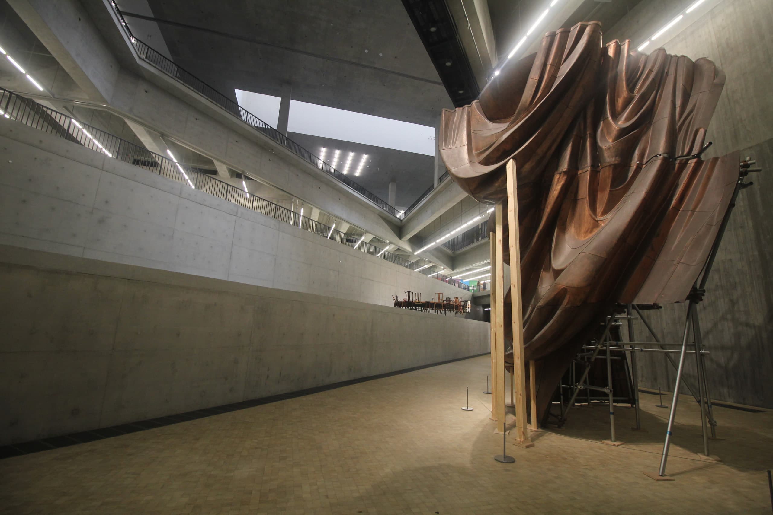 Monumental copper-fold installation rising through the concrete Found Space atrium at M+, West Kowloon