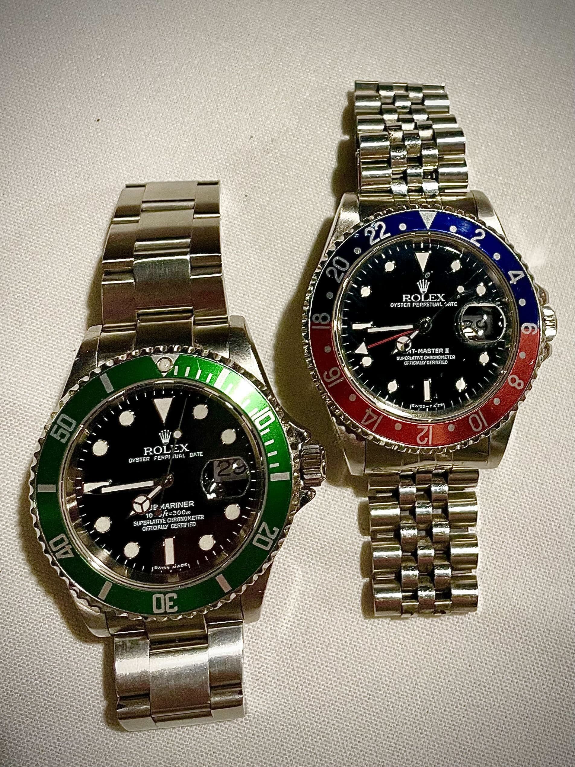A green-bezel Rolex Submariner beside a red-and-blue Rolex GMT-Master II on a pale cloth
