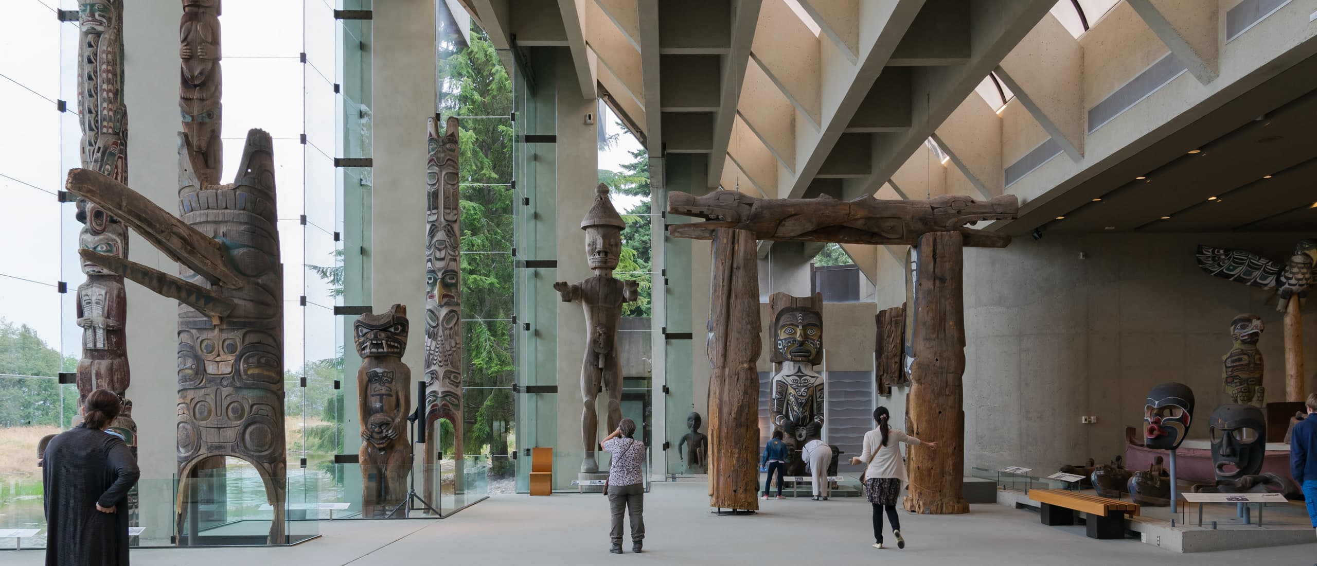 Totem poles and carved house posts in the Great Hall of the Museum of Anthropology at UBC, framed by floor-to-ceiling glass with the North Shore mountains beyond