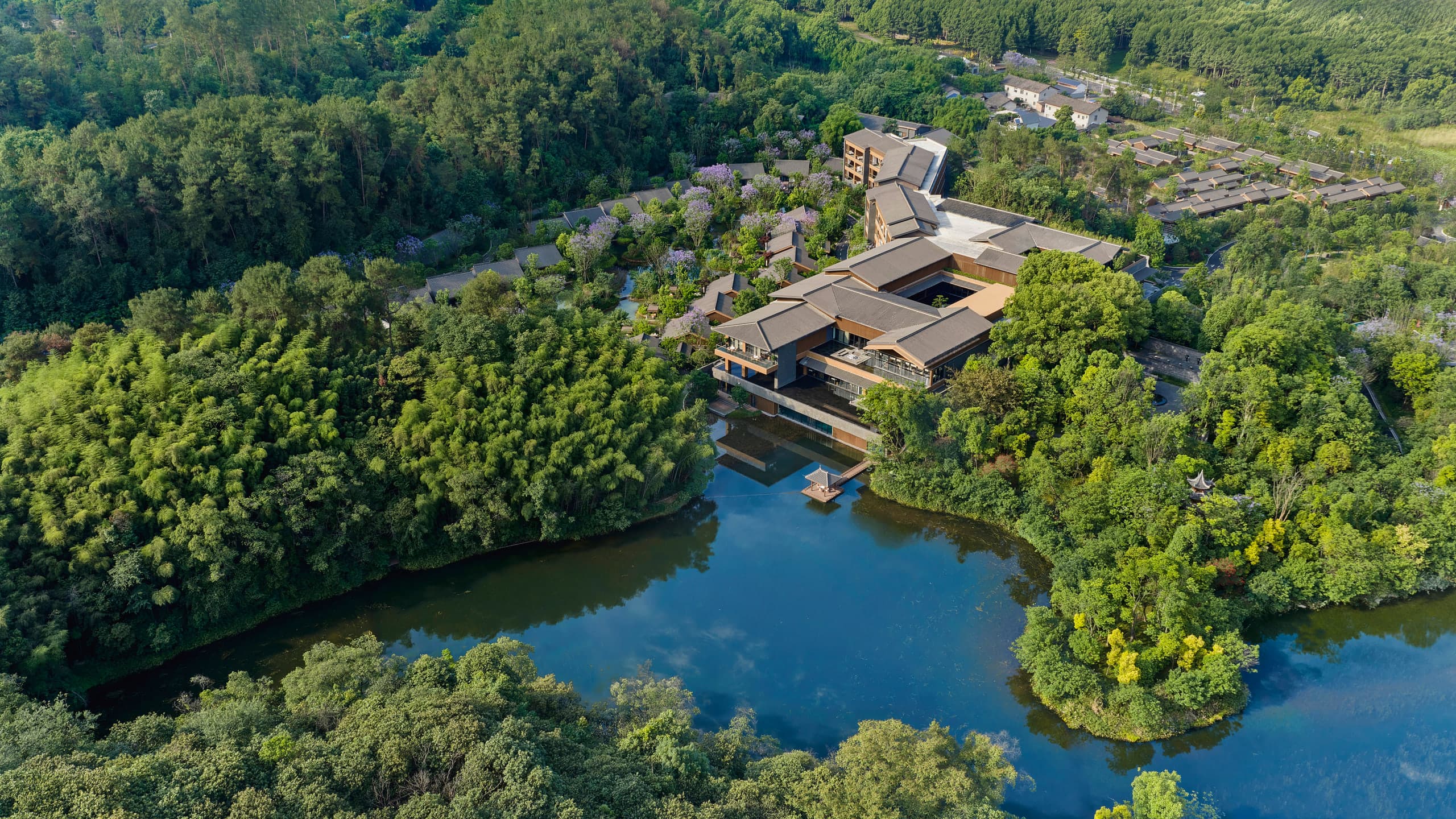 Aerial view of Banyan Tree Mount Emei: a low courtyard lodge beside a lake, with villa roofs in bamboo forest