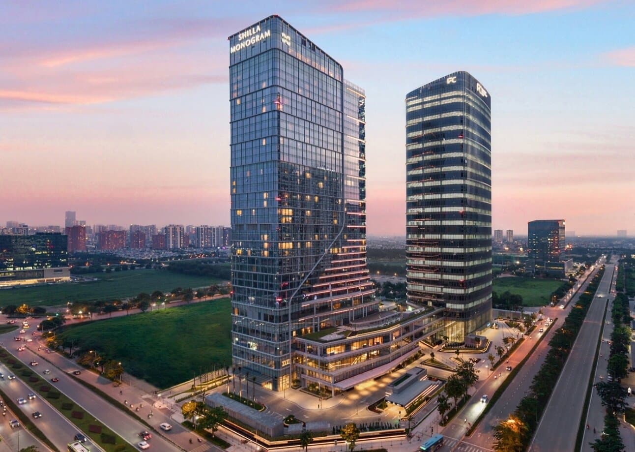 Dusk view of the glass Shilla Monogram tower in Starlake, Hanoi, with the name lit on the crown and a neighboring office tower beside the road