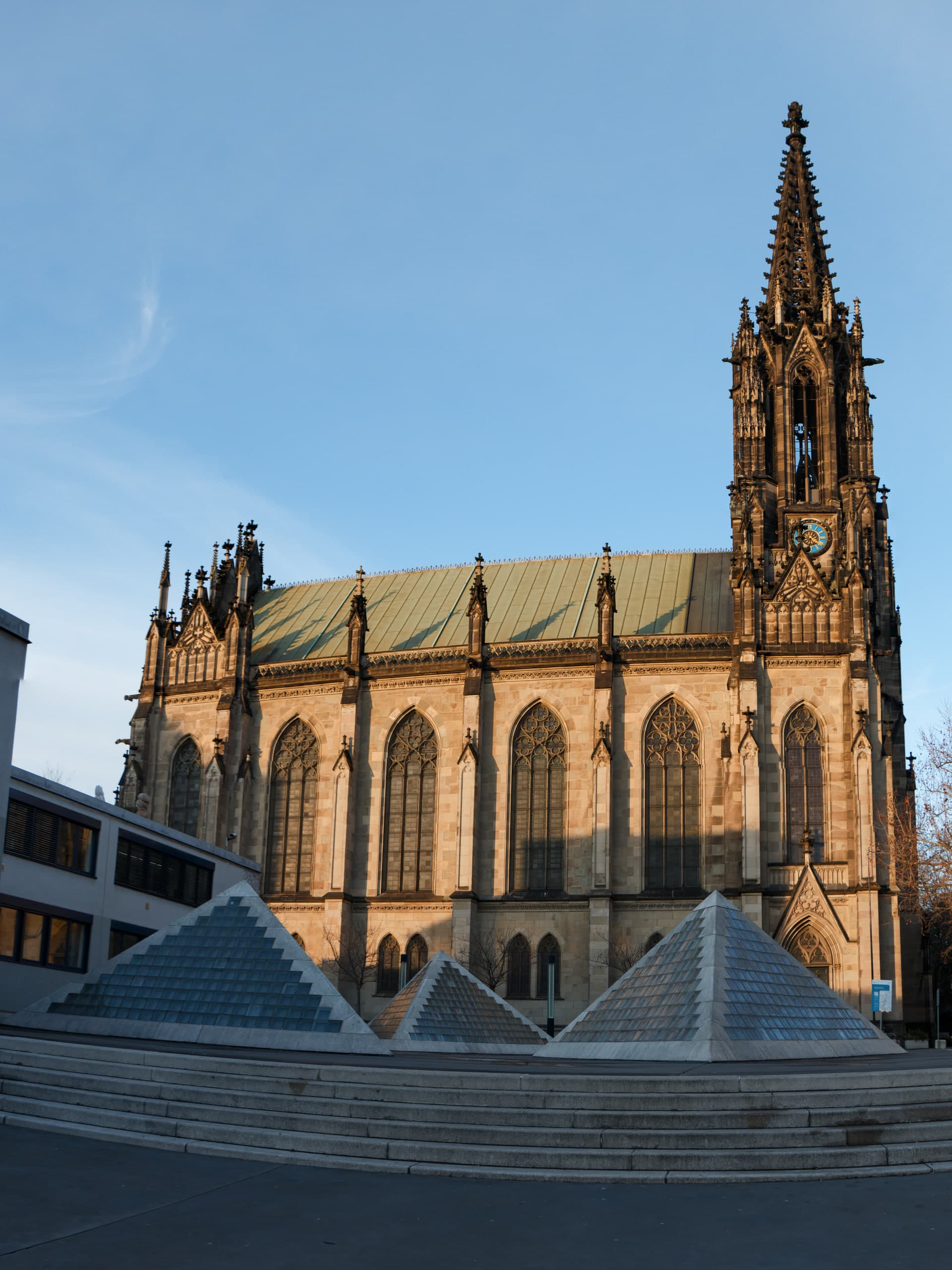 Neo-Gothic Elisabethenkirche in Basel at golden hour, with glass pyramids in the plaza foreground
