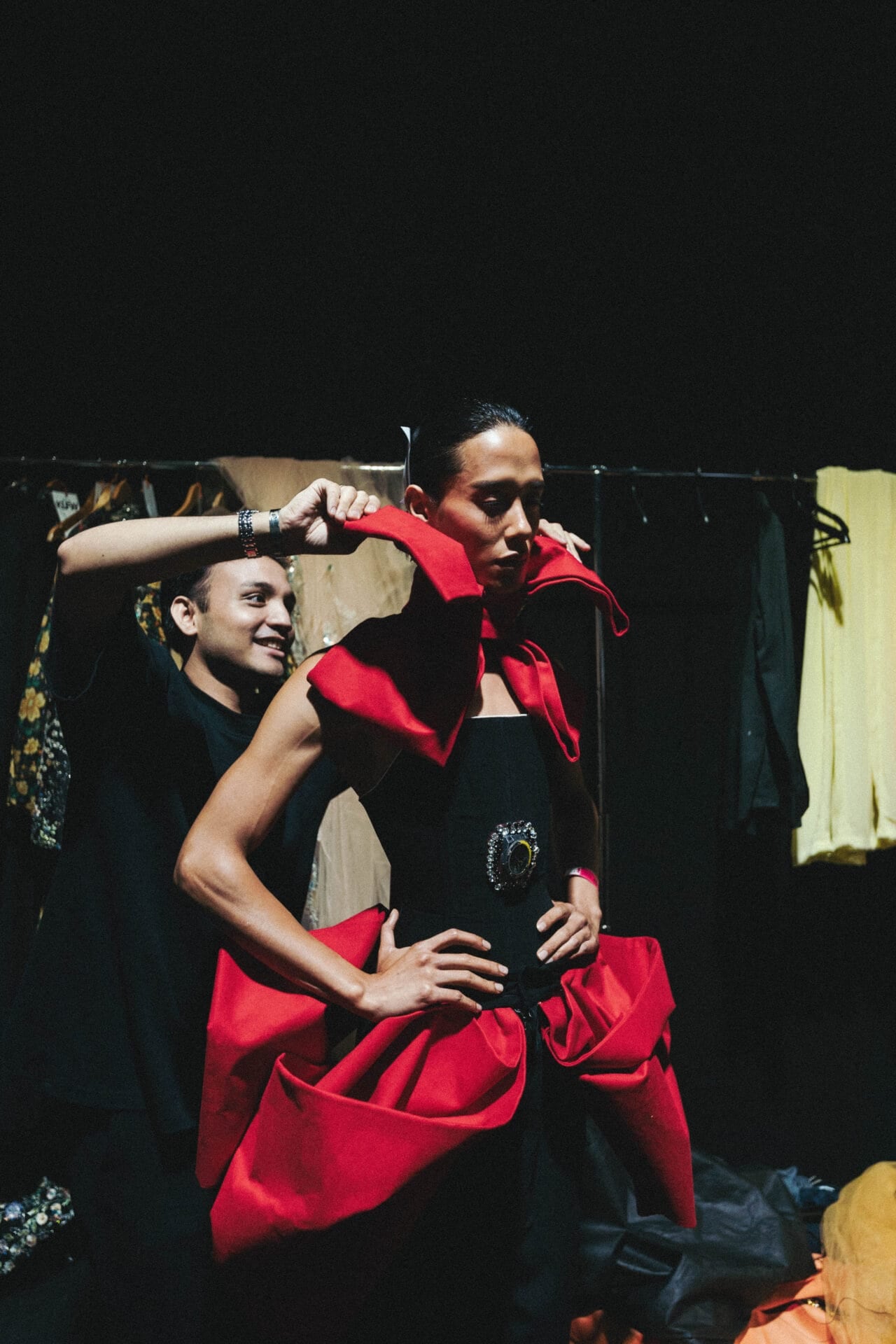 Backstage at KLFW 2026, a model in a black base with a stiff red sculptural collar being adjusted