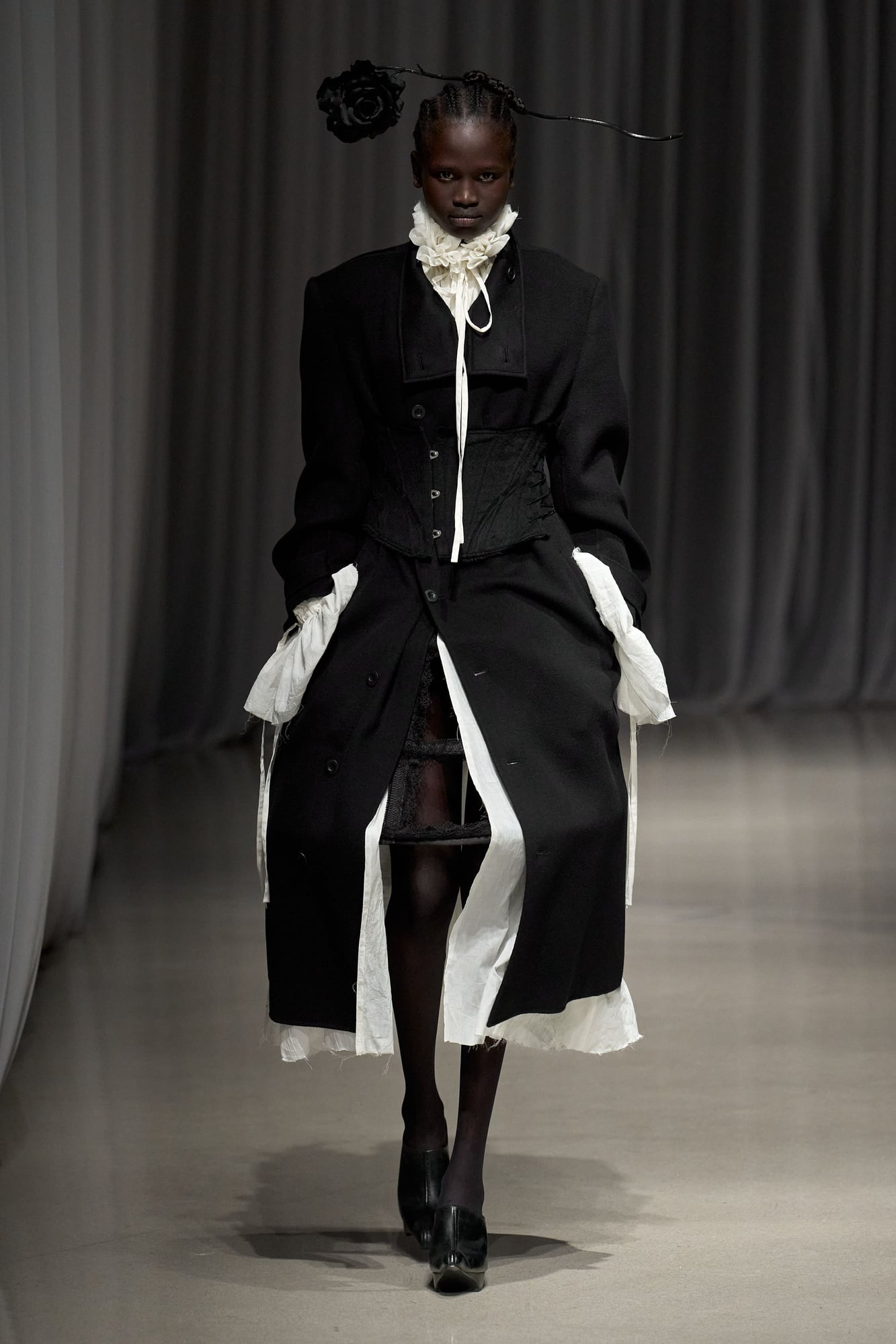 Black coat cinched with a corset over white ruffles, with pannier structure visible under sheer layers, Mukcyen Fall 2026
