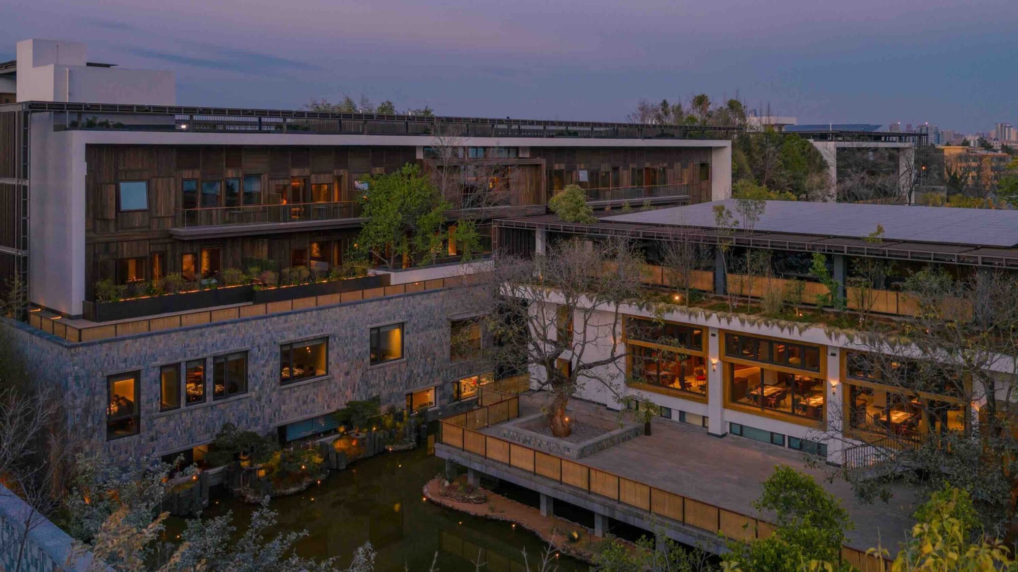 Dusk courtyard at Songtsam Linka Kunming with lit stone-and-timber wings, wooden terrace, and a bare tree over a reflecting pond