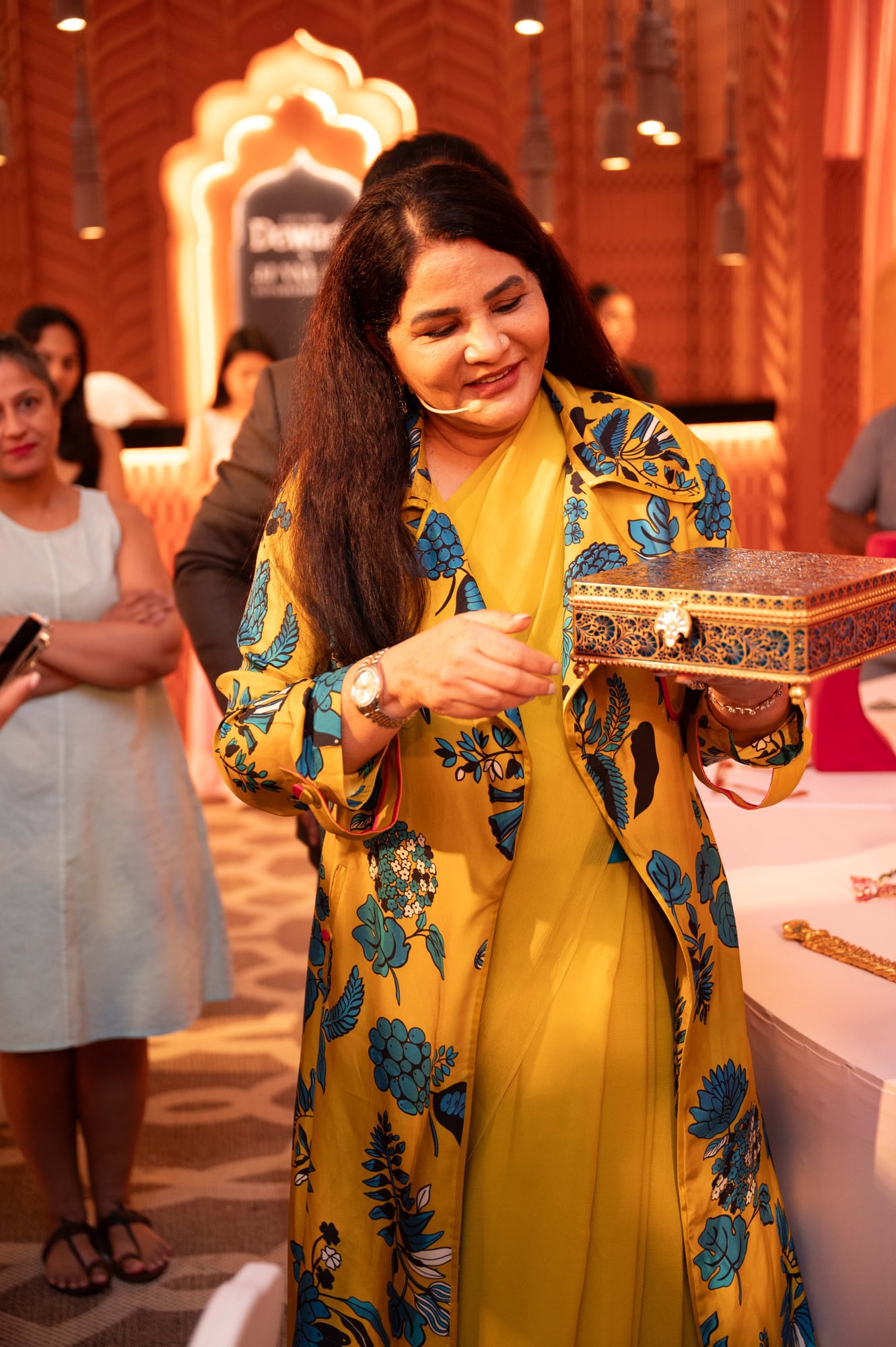 Presenter in mustard sari holding an ornate box beside a high-jewellery display table at Vogue Wedding Atelier, Taj Palace